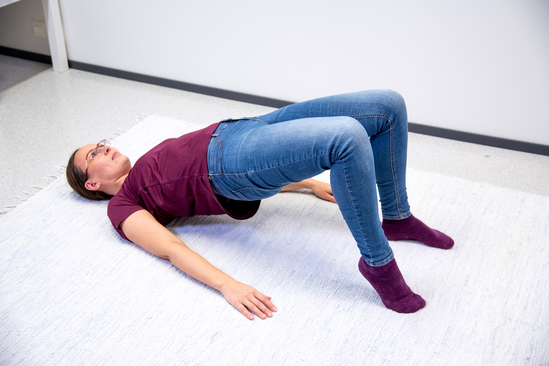 Kvinnan ligger på rygg på golvet med knäna böjda och fotsulorna i golvet. Hon har lyft höften och höger häl upp från golvet.