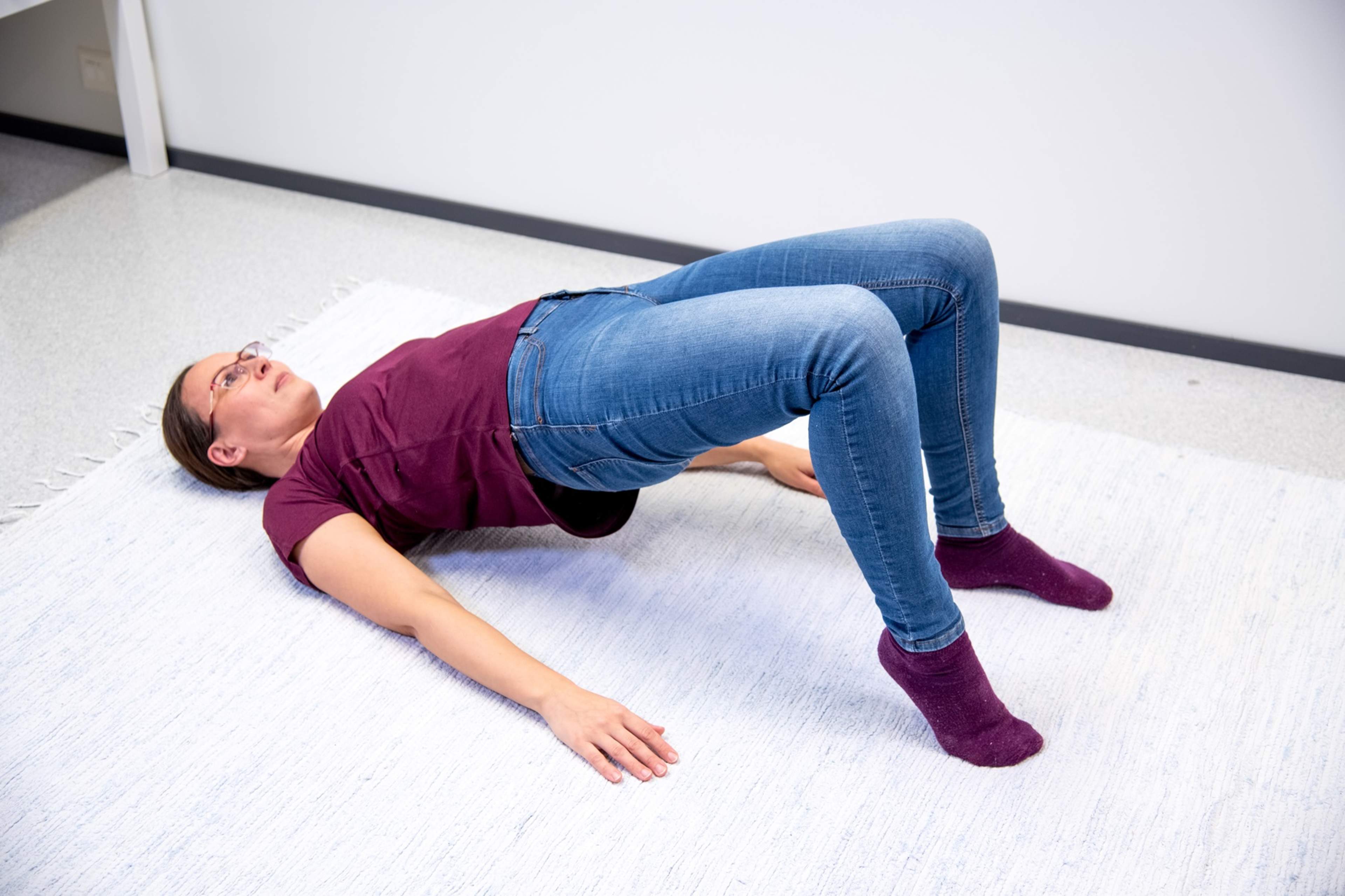 Kvinnan ligger på rygg på golvet med knäna böjda och fotsulorna i golvet. Hon har lyft höften och höger häl upp från golvet.