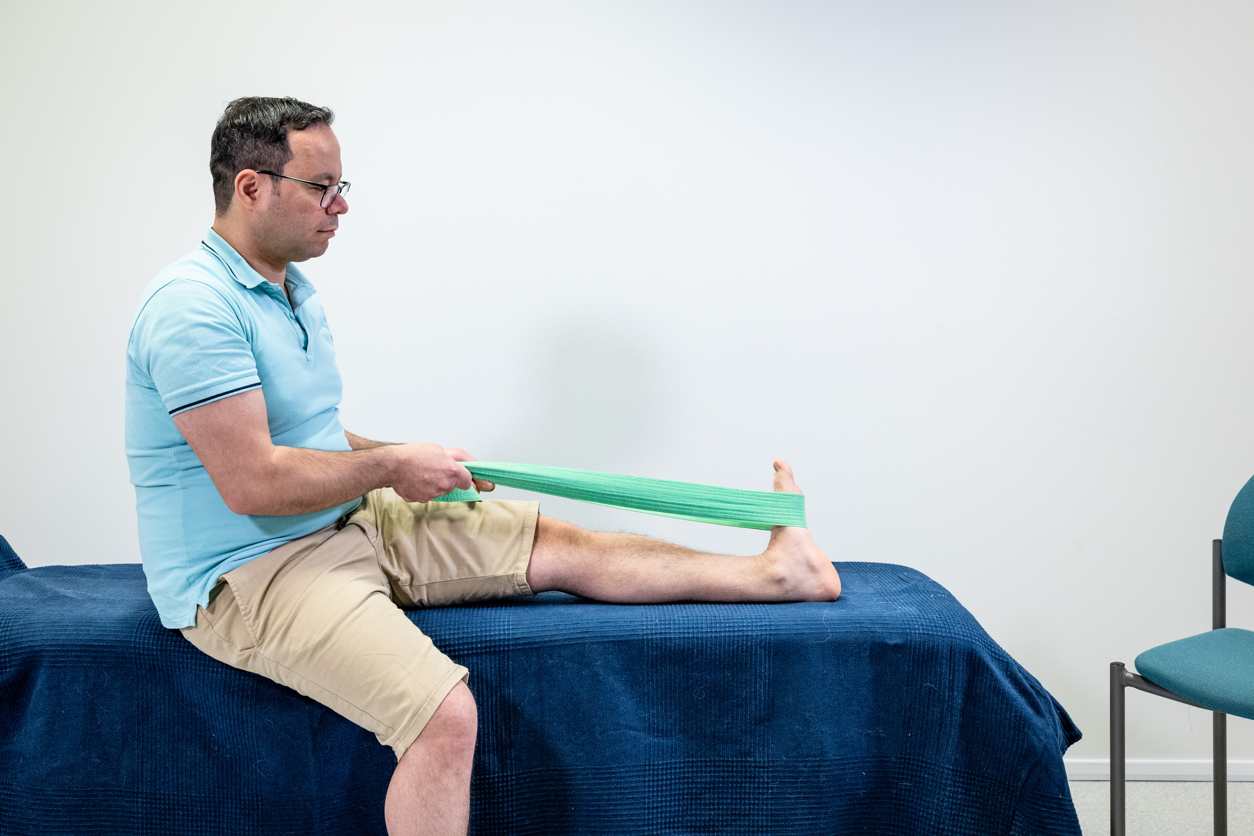 En man sitter på sängen, med vänster ben sträckt rakt ut på sängen och höger ben på golvet. Mannen håller i ett motståndsband som är runt foten. Han har böjt fotleden genom att dra i motståndsbandet.