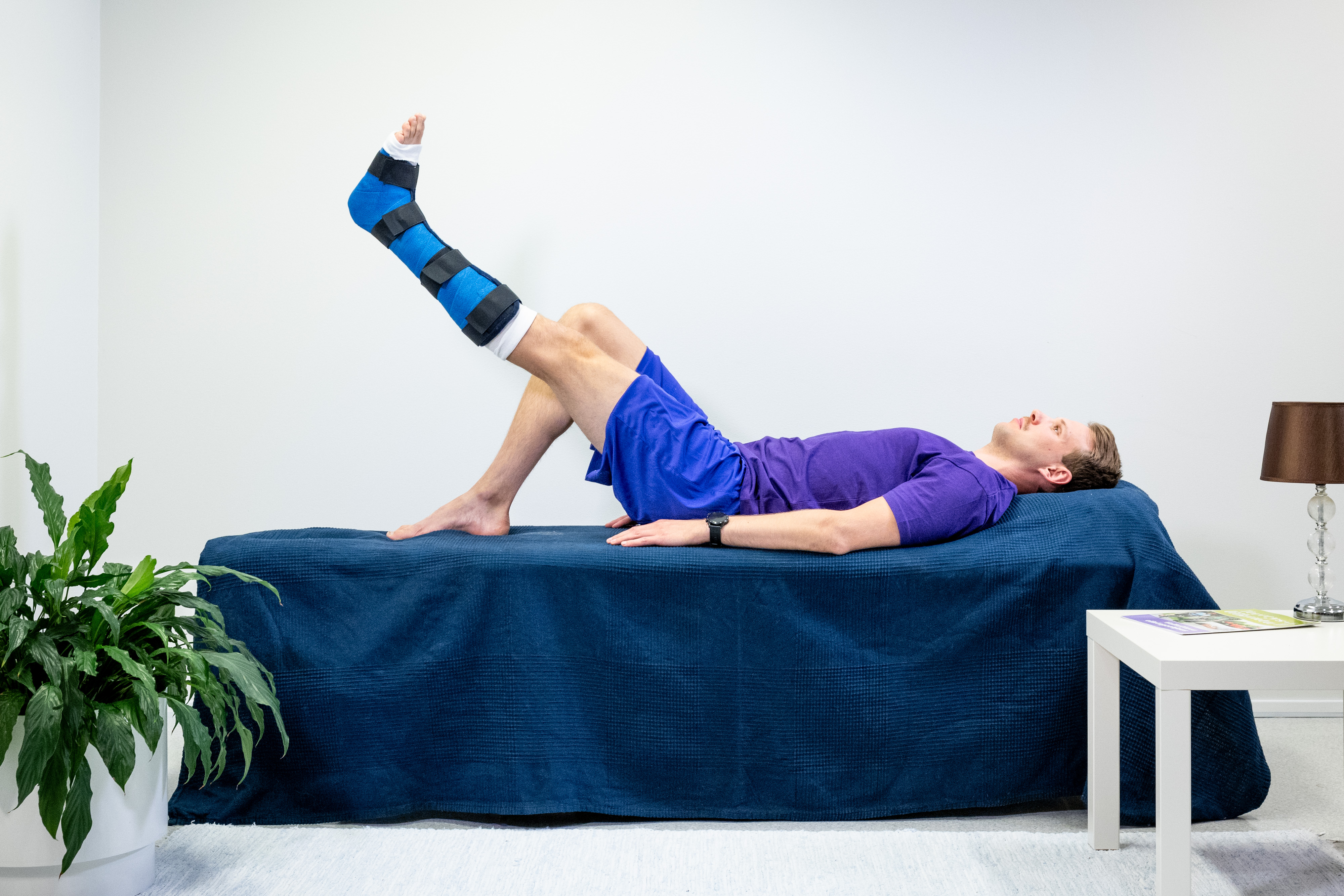 Mannen ligger på rygg i sängen, med ett gips på vänster fotled och höger ben böjt på sängen. Mannen har lyft upp vänster ben så att det är i nivå med knäet på det böjda benet.