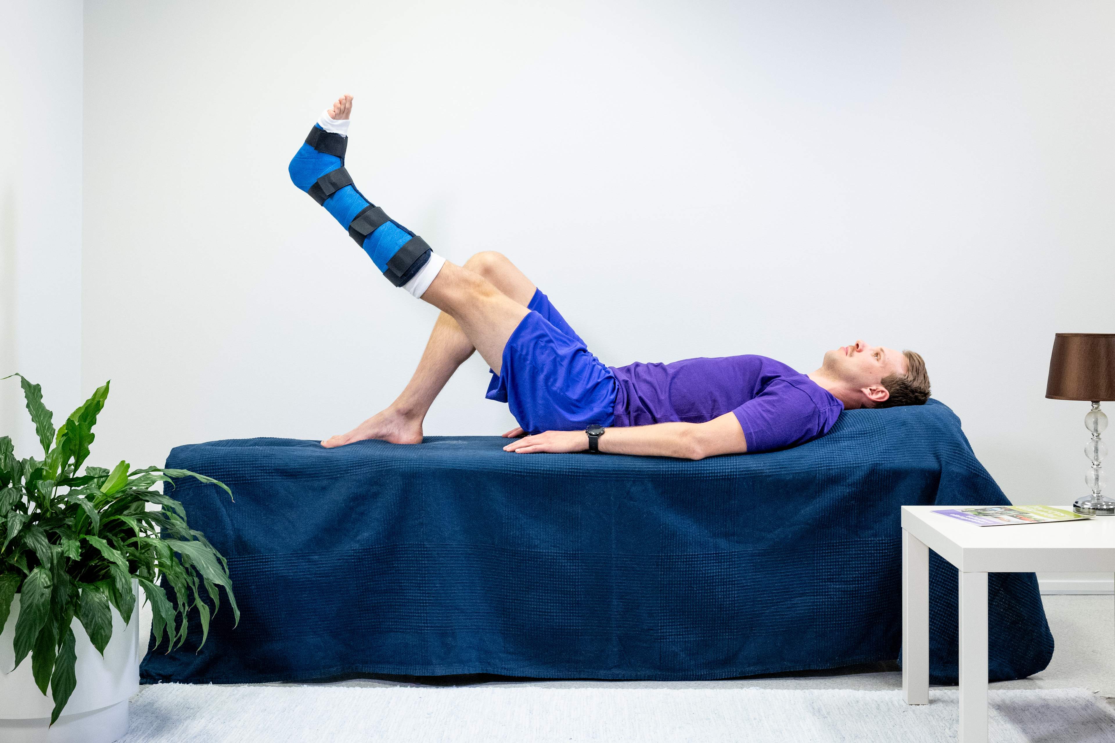 Mannen ligger på rygg i sängen, med ett gips på vänster fotled och höger ben böjt på sängen. Mannen har lyft upp vänster ben så att det är i nivå med knäet på det böjda benet.