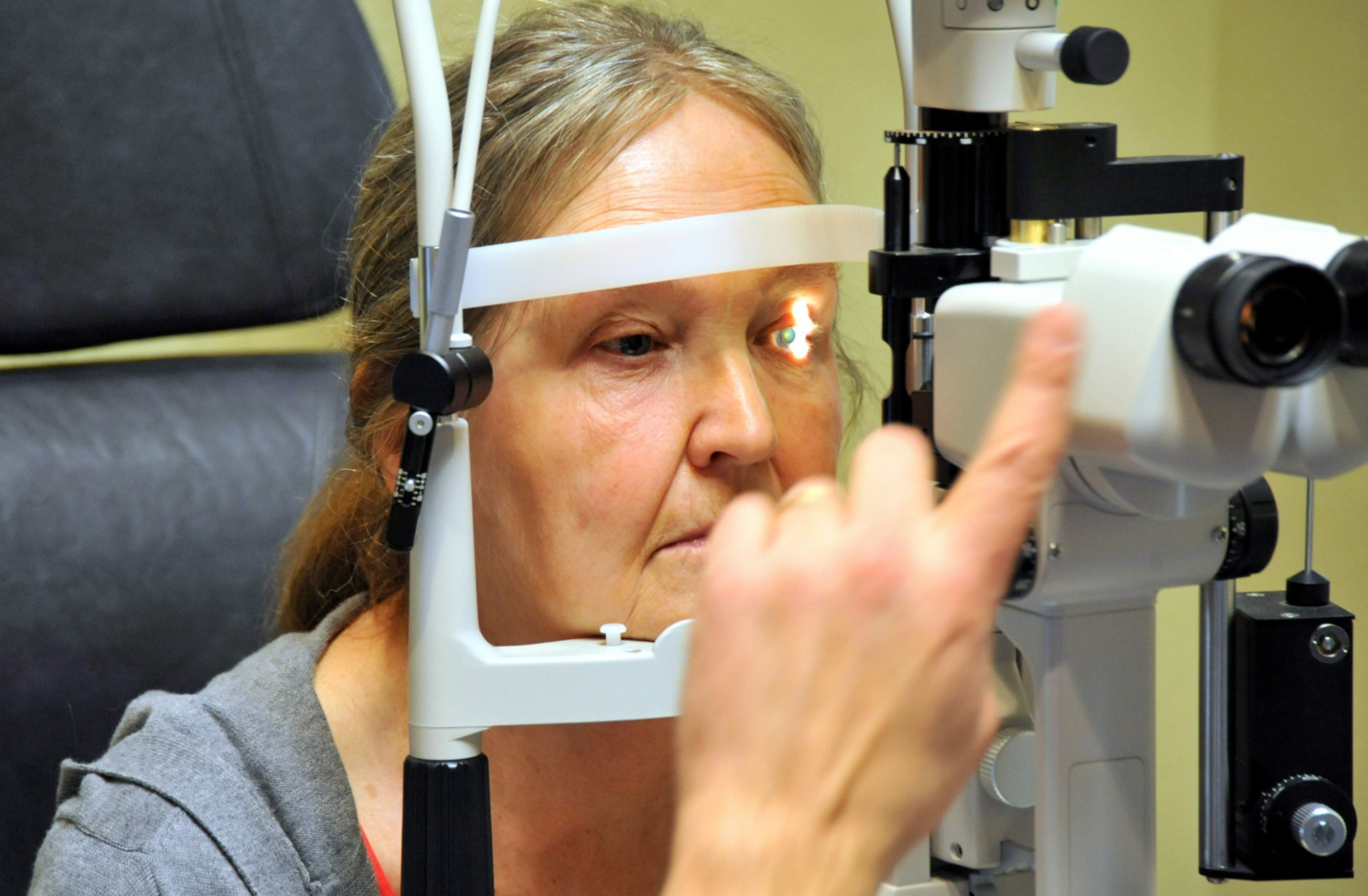 A woman is sat in an examination chair, in front of her is a large white examination device directing light into the eye. In the foreground of the photo are microscope lenses and in front of the device is the hand of the person doing the examination.