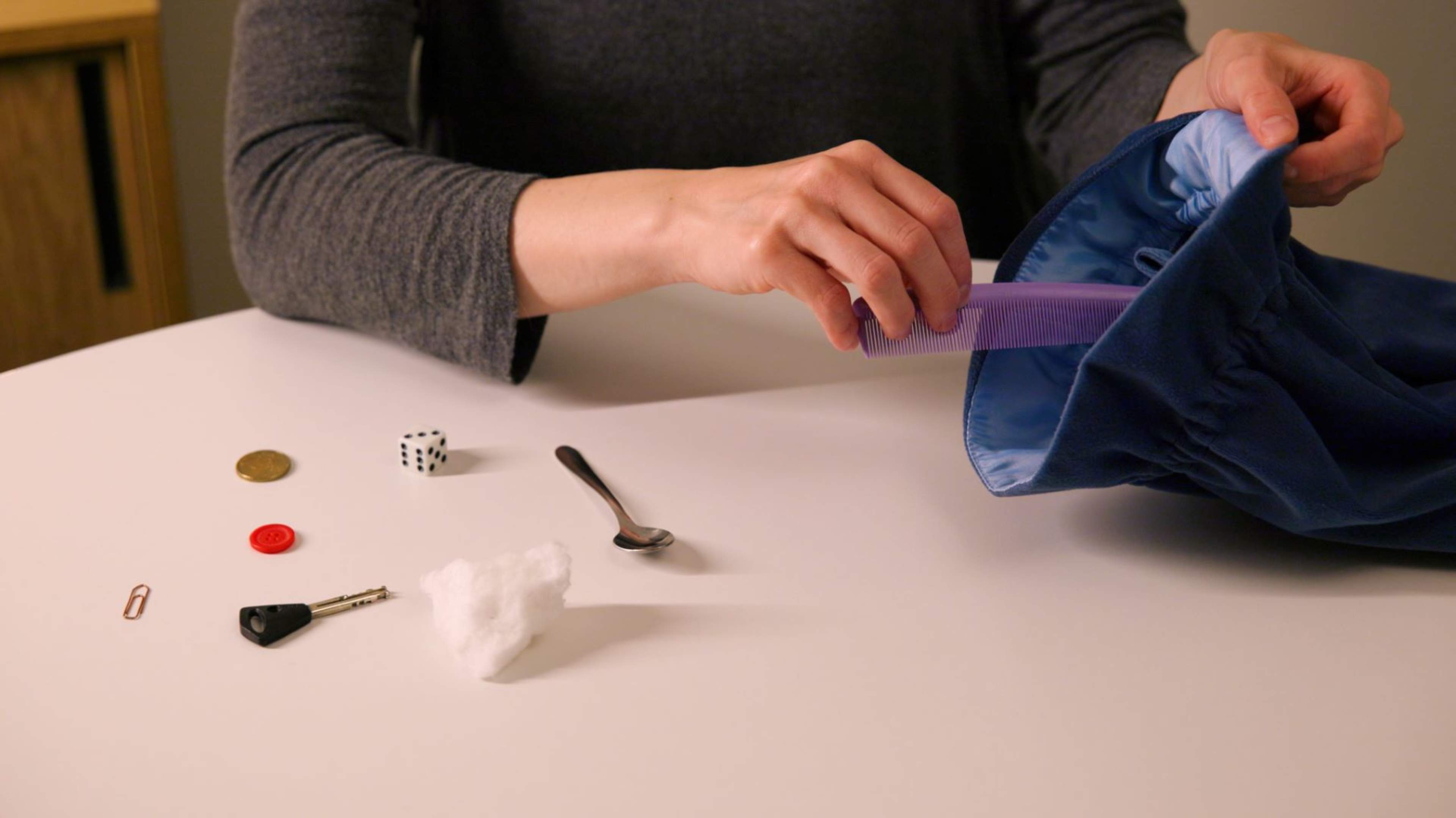 Pöydällä on esineitä kuten lusikka, avain, noppa ja nappi. Henkilöllö on kädessä kampa, jonka hän laittaa pussiin.