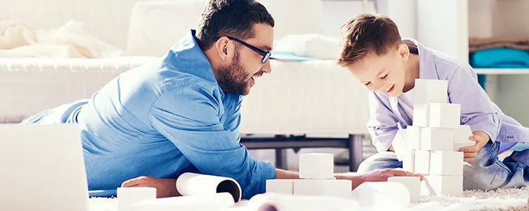 Vater und Sohn spielen mit weißen Bauklötzen auf einem weißen Wohnzimmerteppich vor einem weißen Sofa.