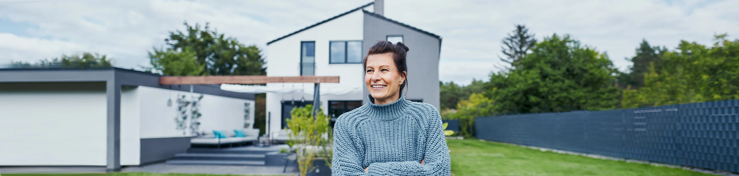 Frau steht im Garten vor Einfamilienhaus und freut sich