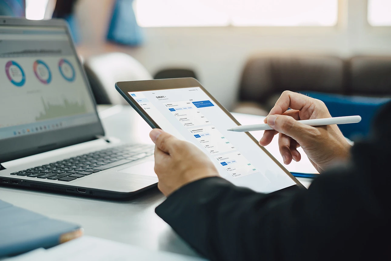 Person analysiert Finanzdaten auf einem Tablet neben einem Laptop im Büro
