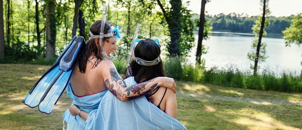 Zwei Personen mit Taucherausrüstung auf grüner Wiese am See