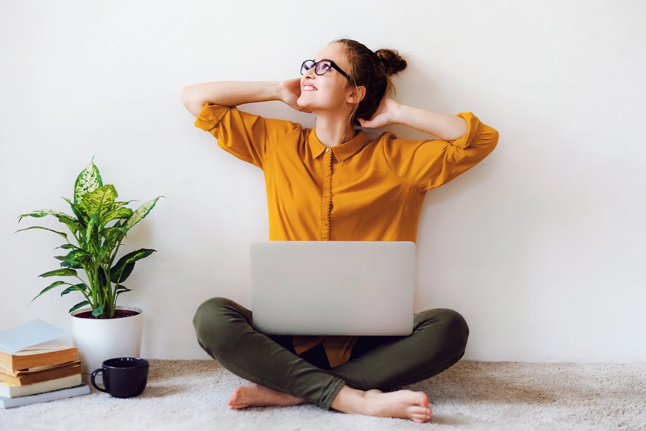 Eine gut gelaunt wirkende Frau sitzt auf dem Boden und hat ihren Laptop auf dem Schoß