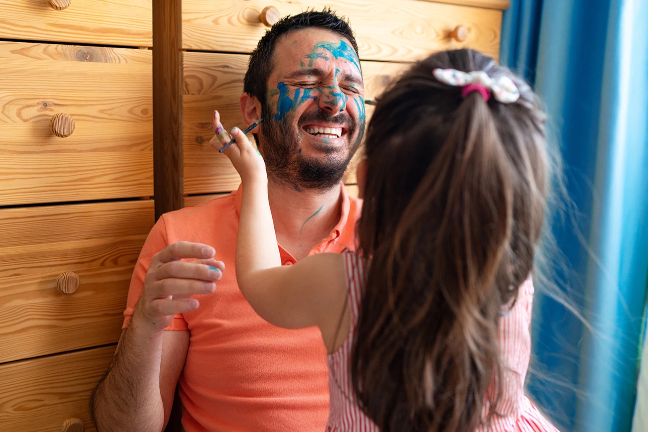 Ein Kind schminkt einen Mann bunt im Gesicht. Beide haben Spaß.