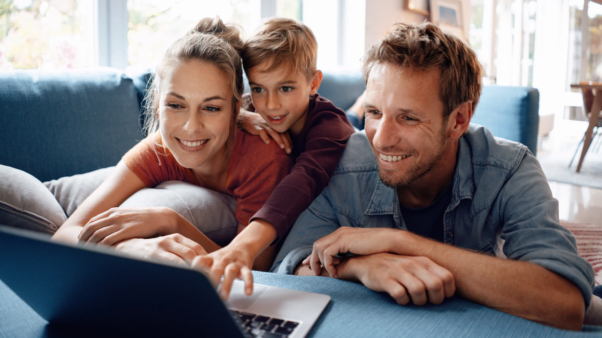 Eine Frau, ein Kind und ein Mann liegen auf der Couch und schauen auf einen Laptop.