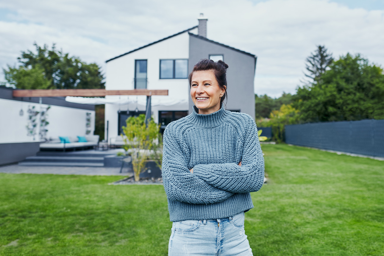 Frau steht im Garten vor Einfamilienhaus und freut sich