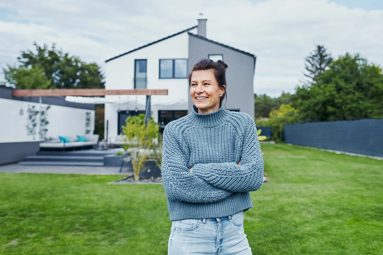 Frau steht im Garten vor Einfamilienhaus und freut sich