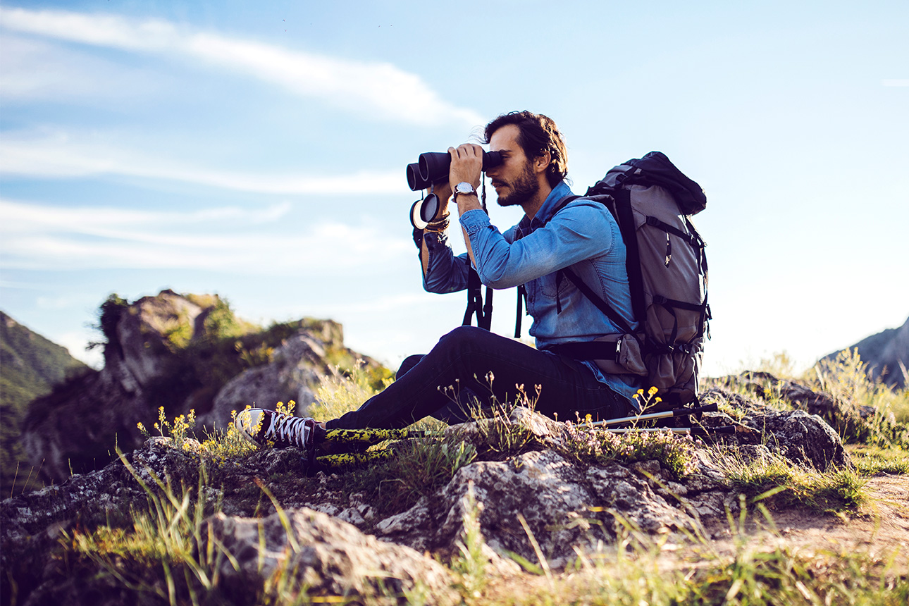 Mann mit Rucksack und Fernglas in Händen sitzt auf Bergfelsen