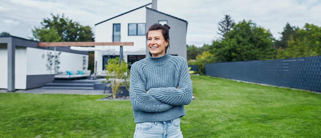 Frau steht im Garten vor Einfamilienhaus und freut sich