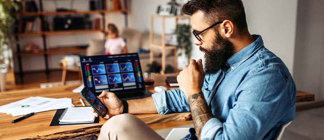 Mann mit Bart sitzt mit Smartphone vor dem Laptop