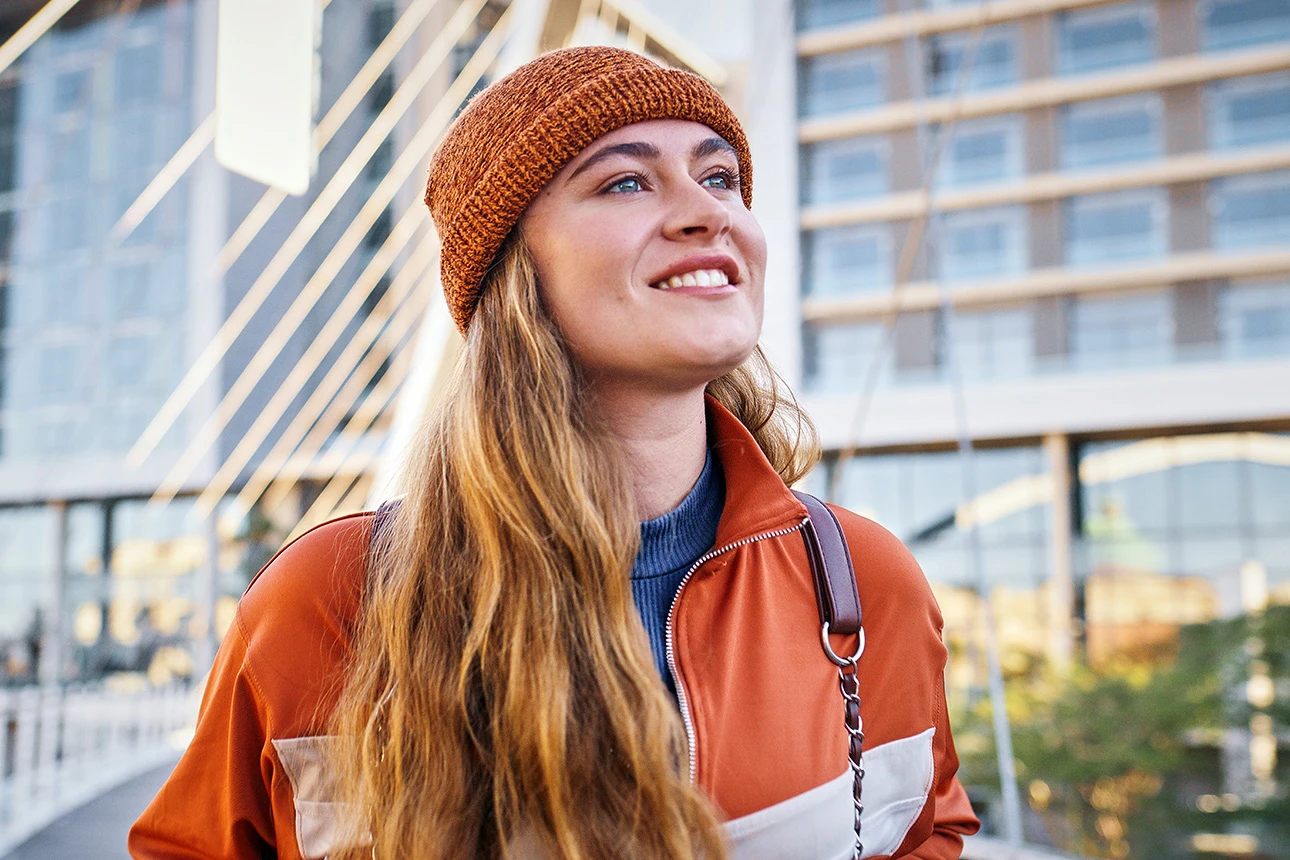 Eine Frau mit Mütze steht vor modernen Gebäuden und lächelt.