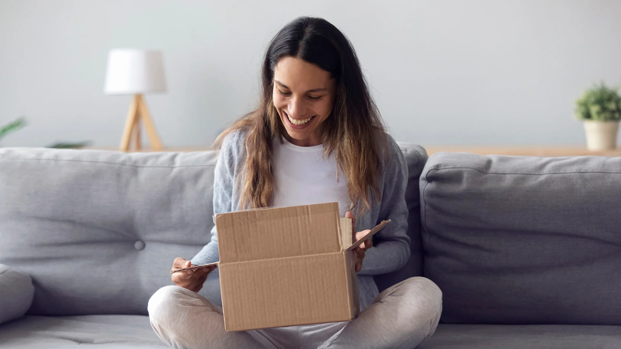 Eine Frau öffnet ein Versandpaket und freut sich während sie auf der Couch sitzt.