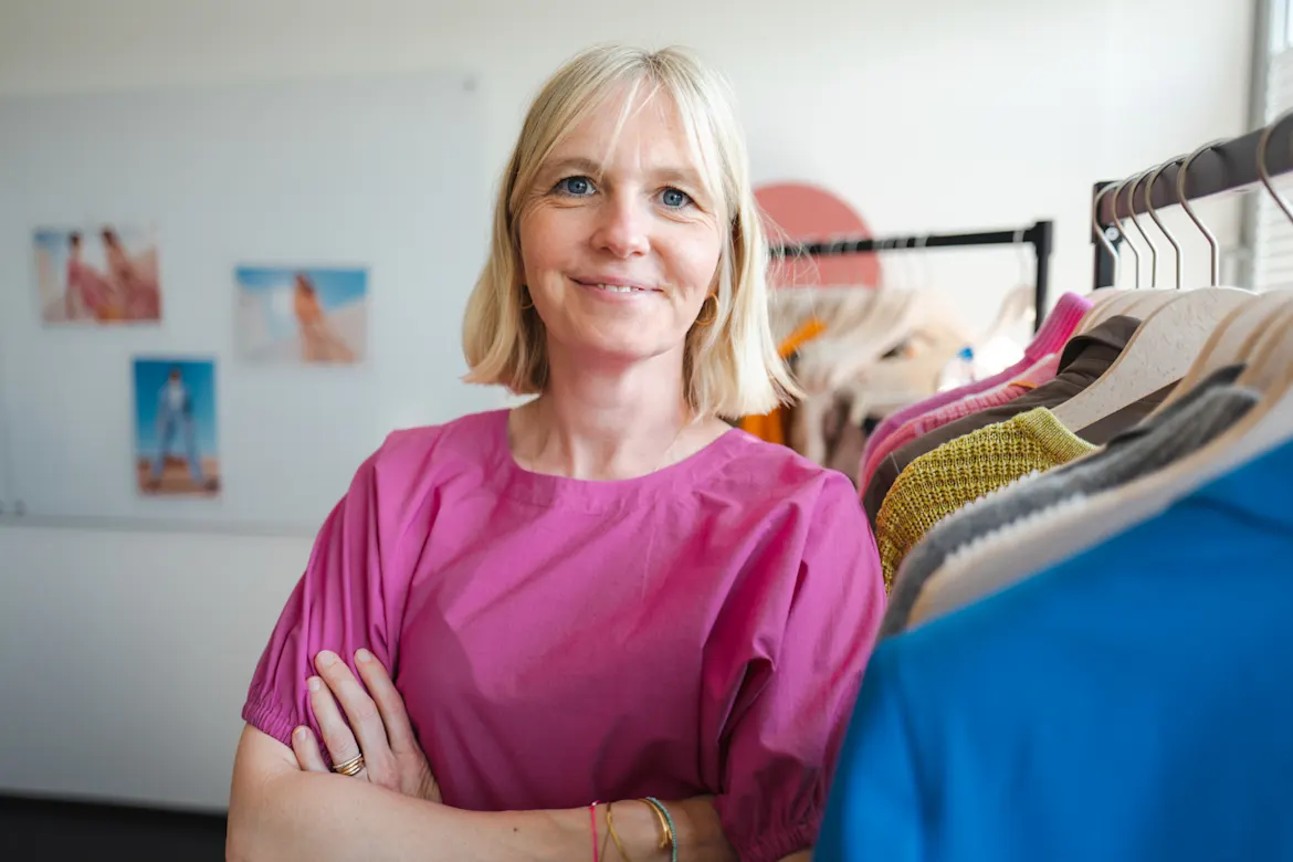 Eine Frau in einem pinken Oberteil steht lächelnd vor einem Kleiderständer mit bunten Kleidungsstücken.