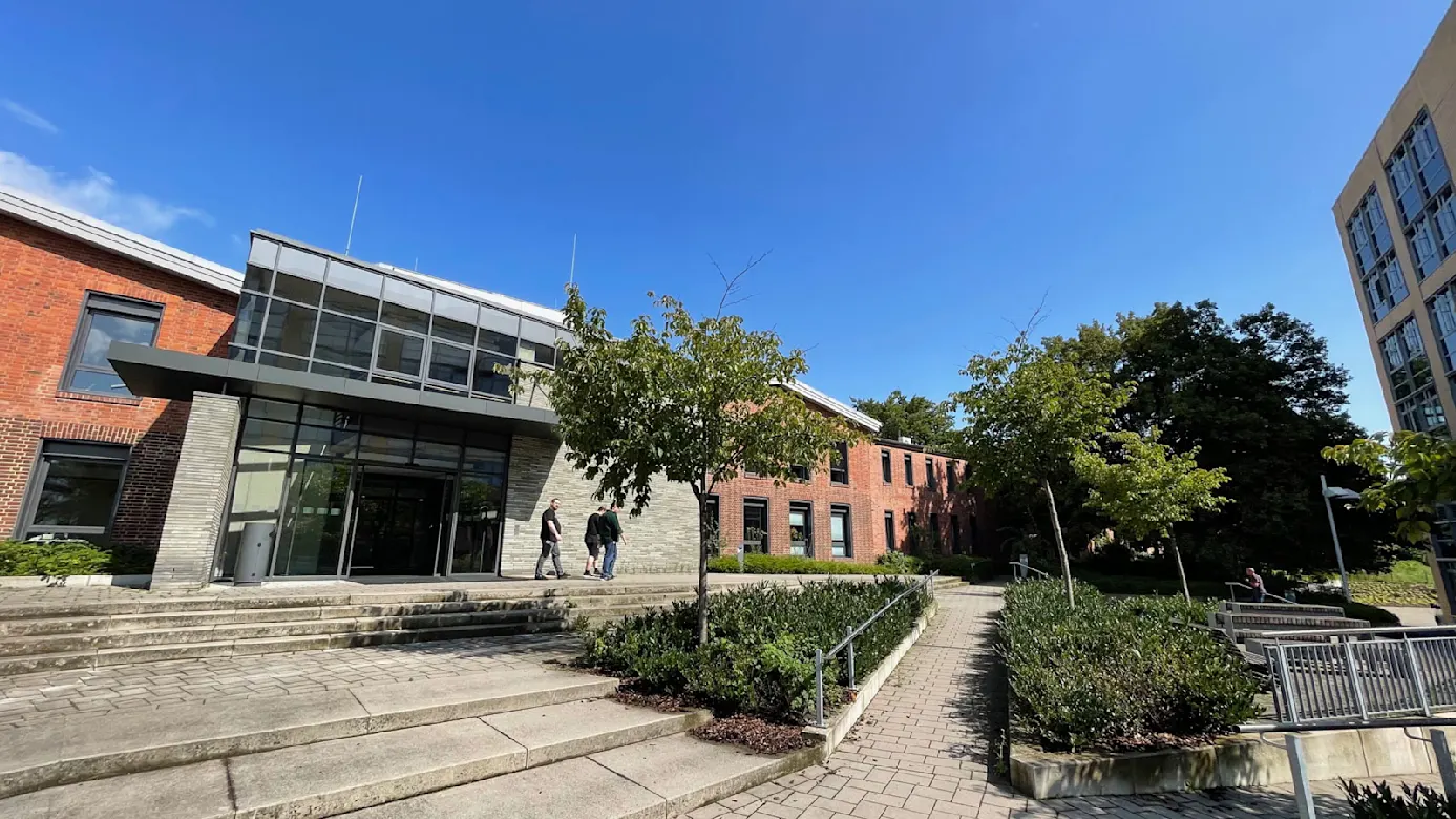 Modernes Universitätsgebäude mit Glasfassade, umgeben von Grünflächen und Bäumen unter blauem Himmel.