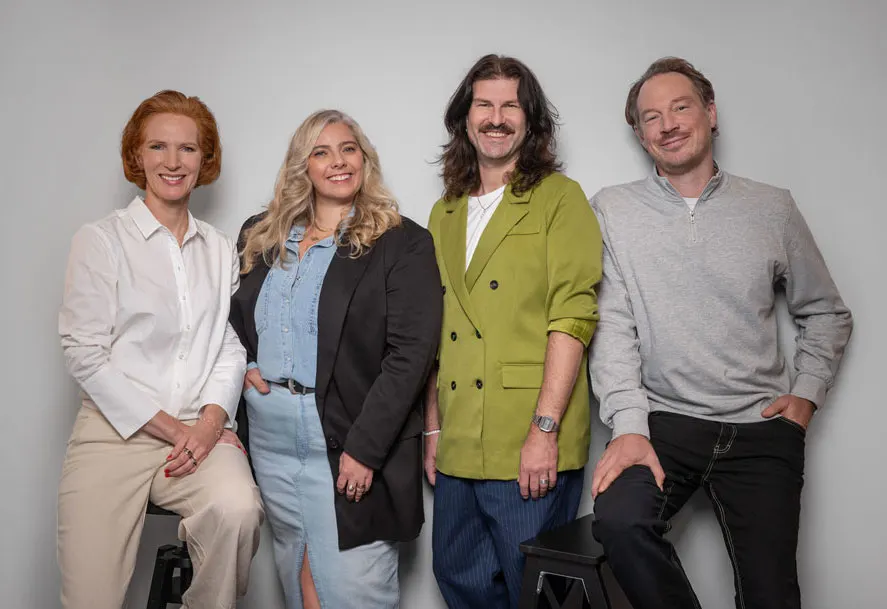 Four smiling people in fashionable clothes stand side by side. The background is neutral.