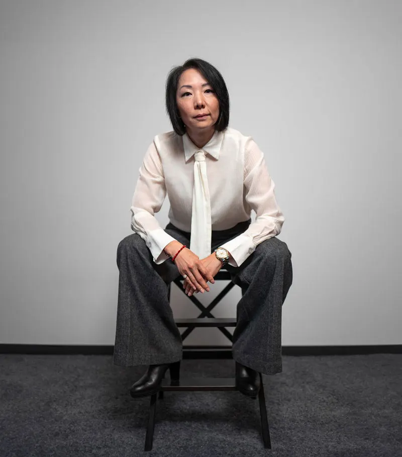 A woman sits hunched in a chair. She has a strong facial expression and is dressed fashionably. The background is neutral.