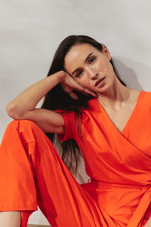 Eine Frau in leuchtendem Orange posiert elegant vor neutralem Hintergrund. Ihr Blick wirkt nachdenklich.