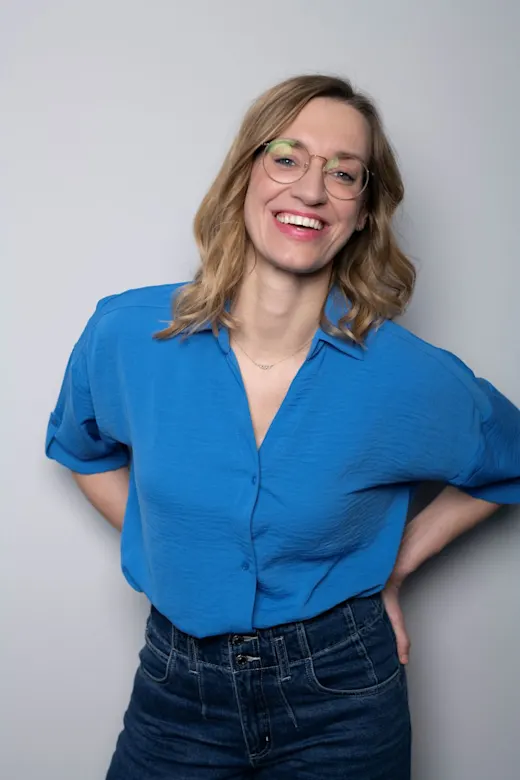 Fröhliche Frau in blauer Bluse und Brille lächelt vor hellem Hintergrund.