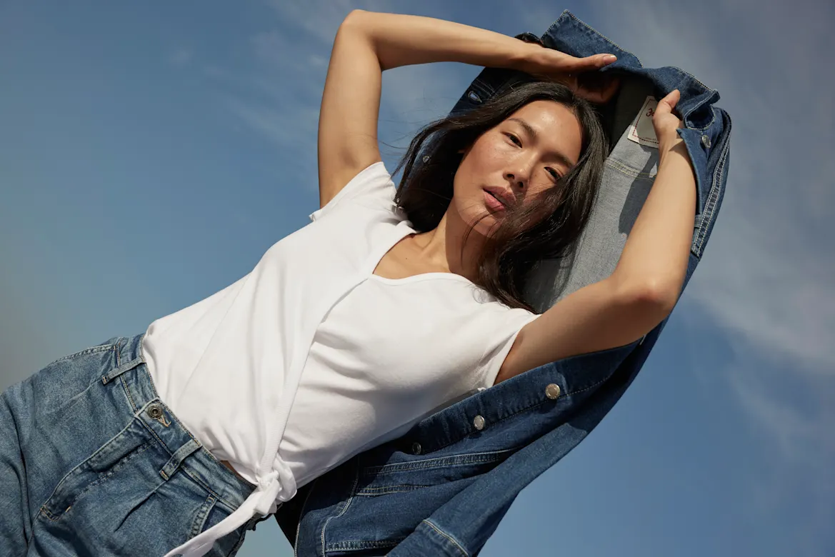 Eine Frau in Jeansjacke und weißem Top posiert selbstbewusst vor blauem Himmel.