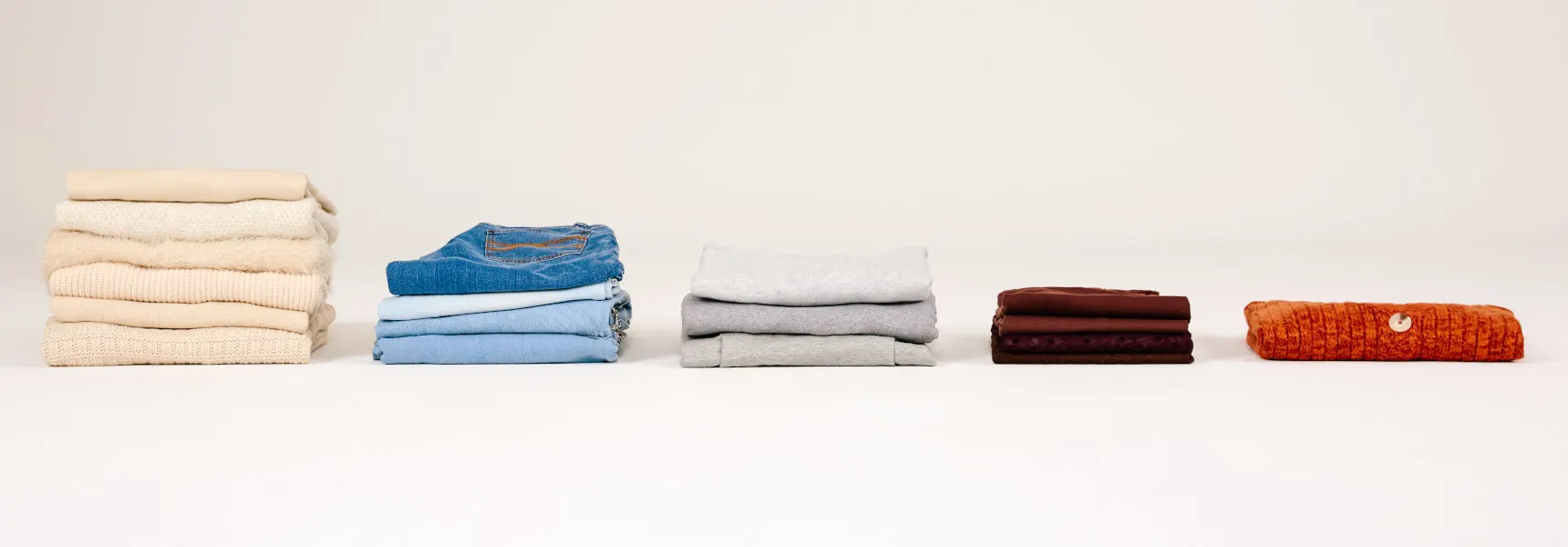 Five piles of clothes lie neatly side-by-side, sorted by color. The piles get smaller from left to right. The background is neutral and simple.