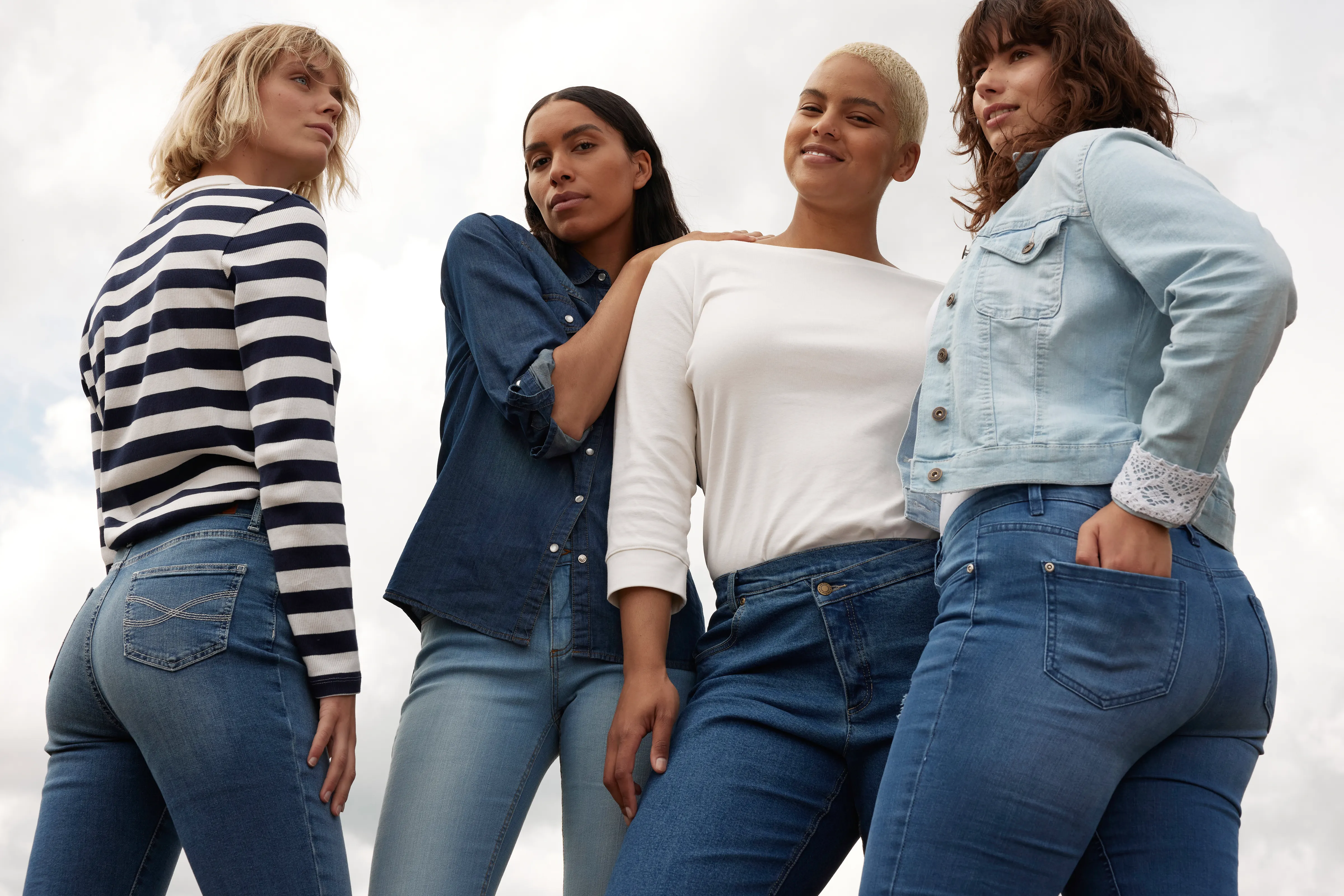 Vier Frauen in Jeans-Outfits posieren selbstbewusst vor hellem Himmel.