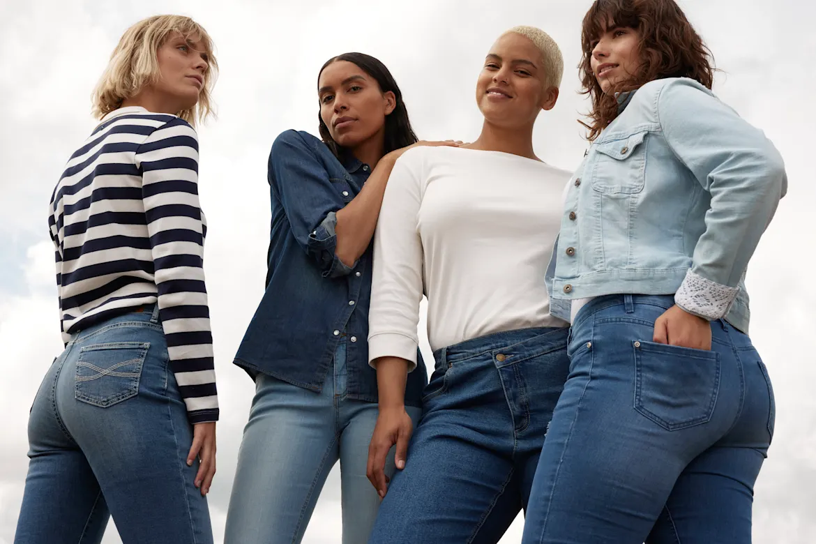 Vier Frauen in Jeans und verschiedenen Oberteilen posieren selbstbewusst nebeneinander.