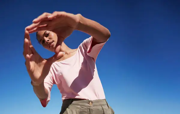 Eine Person in rosa Shirt vor blauem Himmel, die Arme schützend über dem Kopf.