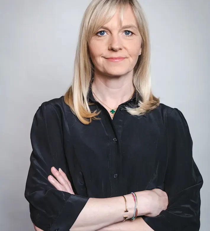 Die Geschäfsführerin Carolin Klar lächelt selbstbewusst mit verschränkten Armen vor grauem Hintergrund.
