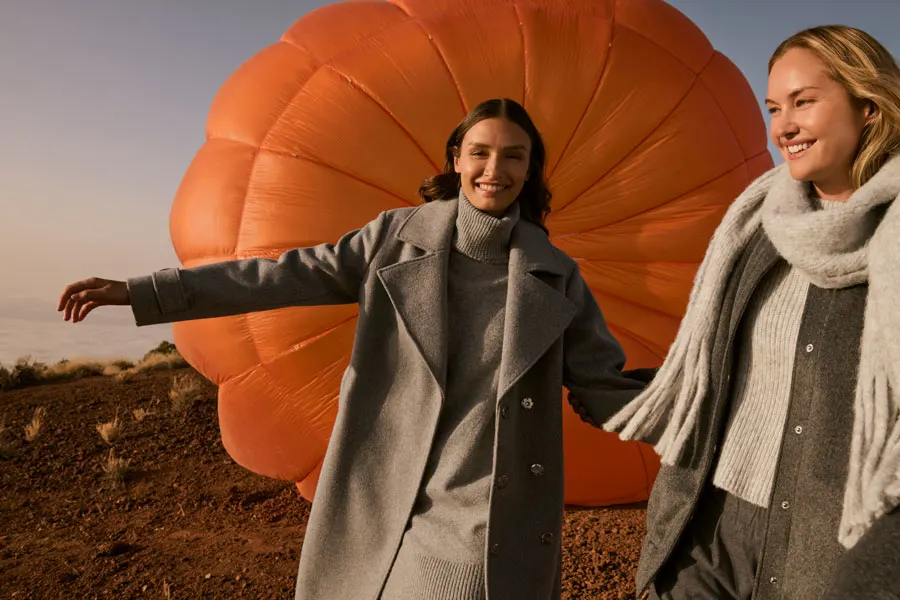 Zwei Frauen stehen nebeneinander vor einem orangefarbenen Heißluftballon. Sie tragen warme Mäntel und lächeln. Der Hintergrund zeigt eine natürliche Landschaft mit klarem Himmel. 