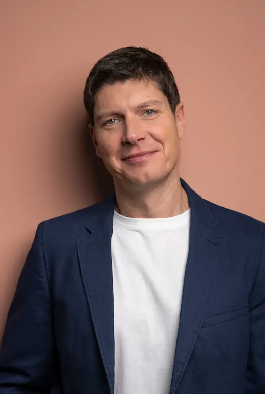 Eine Person in blauem Blazer und weißem T-Shirt vor einem rosa Hintergrund lächelt freundlich.