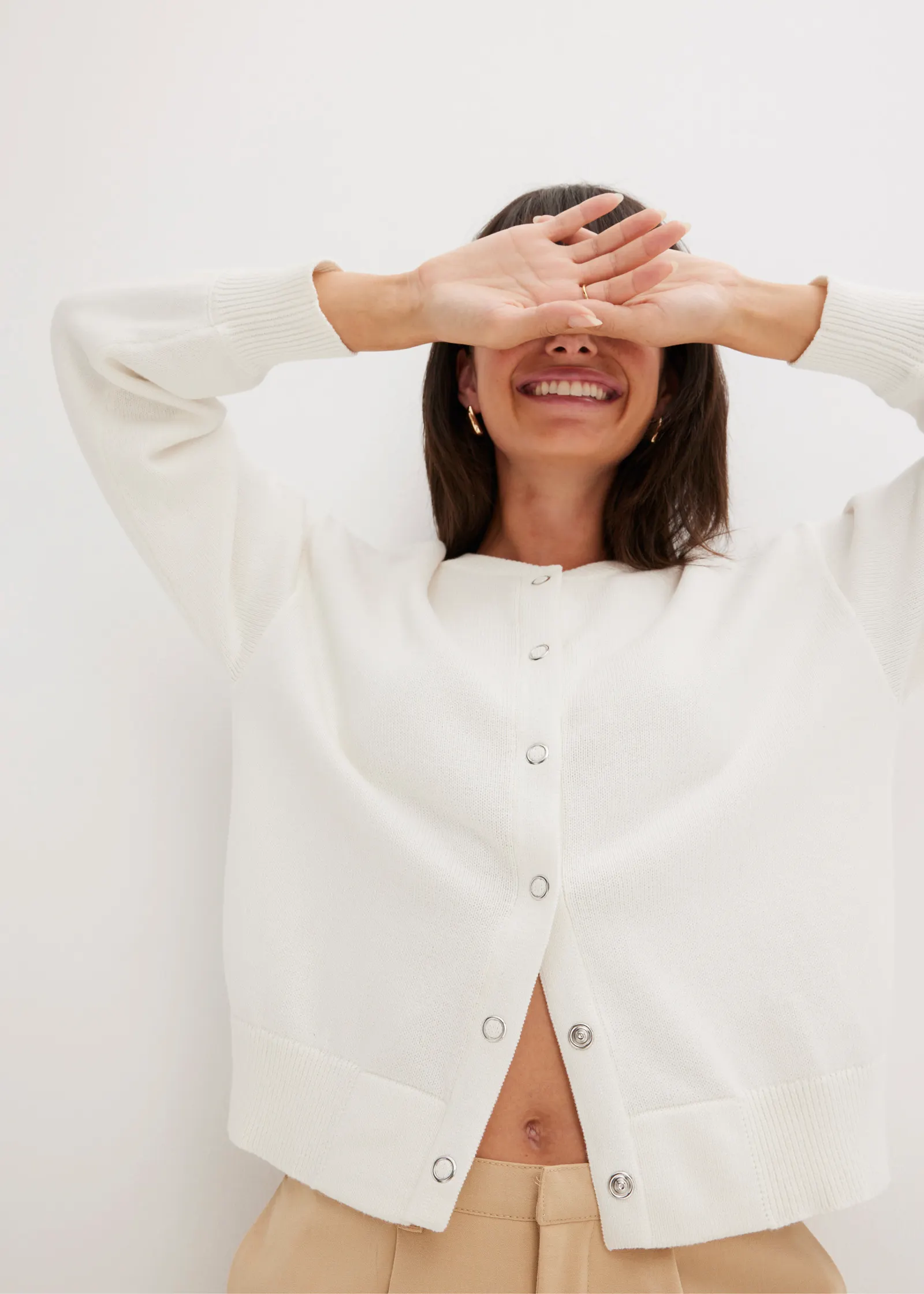 Eine Frau steht vor einer hellen Wand und hält sich die Handrücken vor die Augen. Sie hat ein breites Lächeln und trägt eine weiße Strickjacke mit Knöpfen.