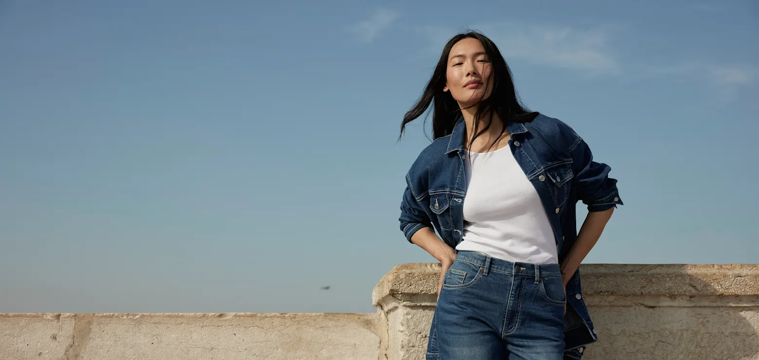 Eine Person in lässiger Jeansjacke und Jeans lehnt an einer Mauer unter blauem Himmel.