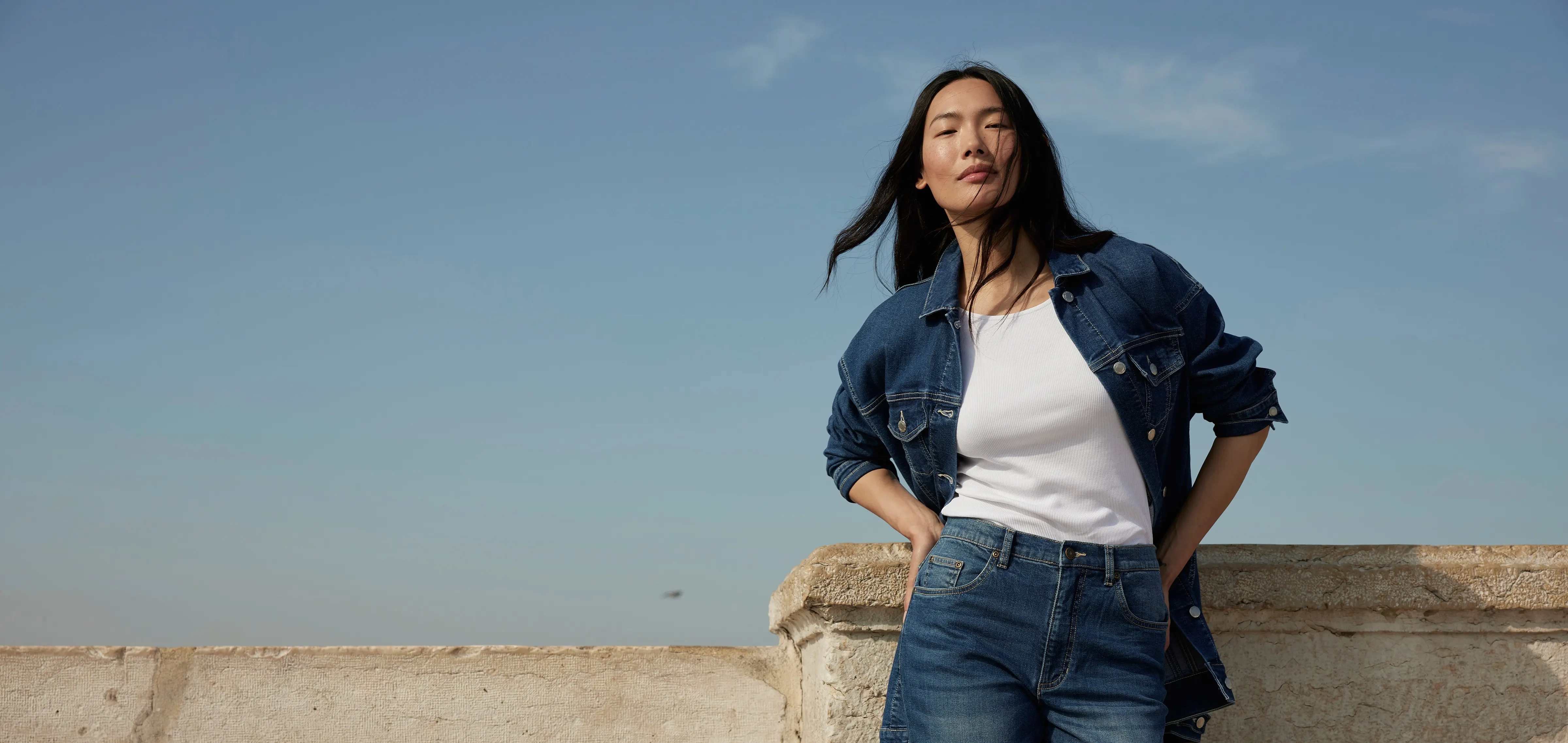 Eine Frau in Denim Look steht vor einer Mauer. Sie stützt ihre Hände auf der Hüfte ab und schaut neutral in die Kamera. Im Hintergrund sieht man den blauen Himmel.