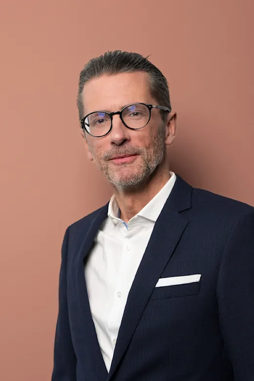 Eleganter reifer Mann mit Brille und Bart im dunklen Anzug vor rosafarbenem Hintergrund.