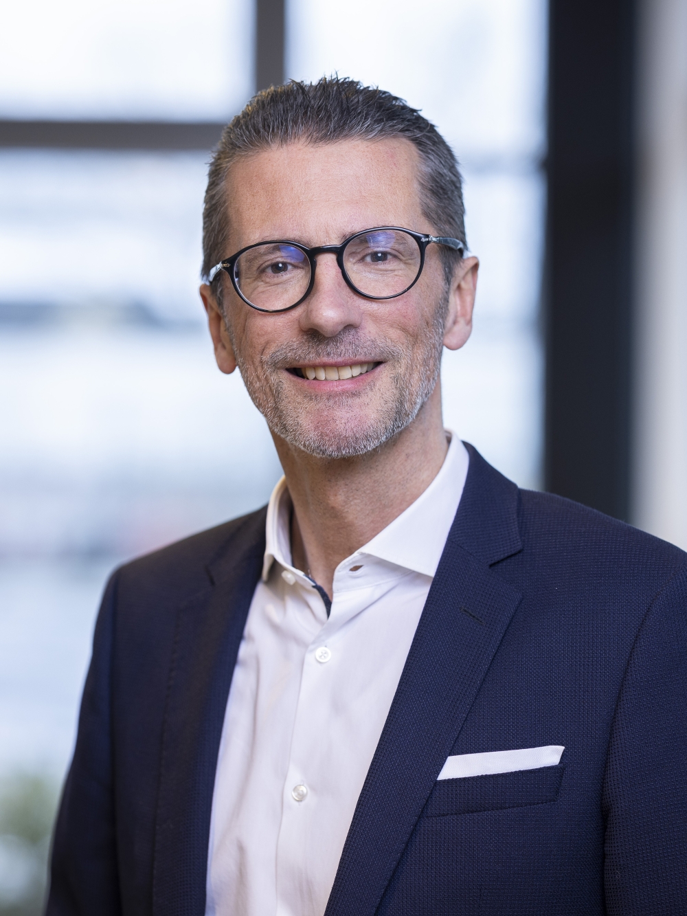 Kai Heck in a dark blue suit and white shirt in front of a bright office window.
