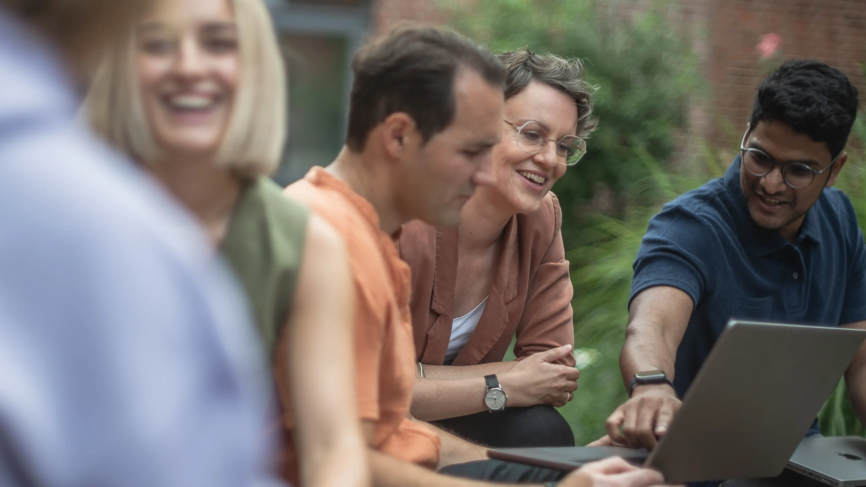 A group of five people sit together outdoors, looking at a laptop. One person points at the screen while the others smile attentively. In the background, green plants and a brick building can be seen.