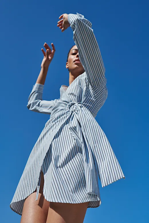 Eine Frau in gestreiftem Hemdkleid posiert dynamisch gegen einen strahlend blauen Himmel.