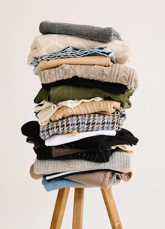 Ein hoher Stapel verschiedener Pullover und Kleidungsstücke liegt ordentlich gefaltet auf einem Holzstuhl.
