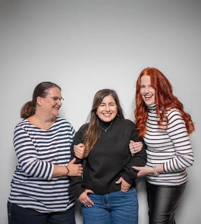 Drei fröhliche Frauen umarmen sich lachend. Sie tragen gestreifte Shirts und einen schwarzen Pullover.