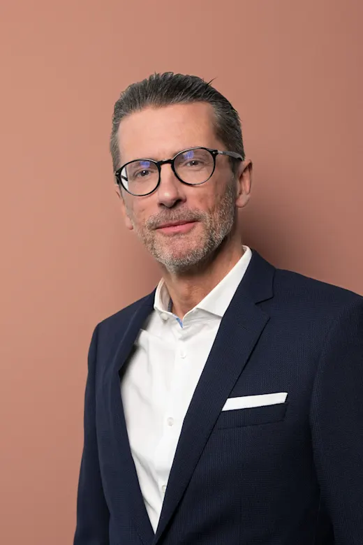 Eleganter Herr mit Brille und Bart trägt dunklen Anzug vor pastellfarbenem Hintergrund.