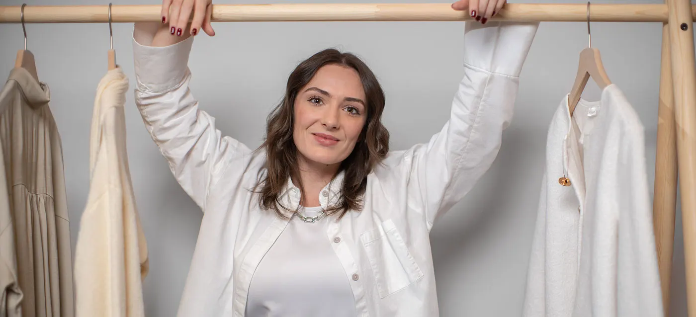 Eine Person in weißer Kleidung streckt sich fröhlich vor einer Kleiderstange mit hellen Kleidungsstücken.
