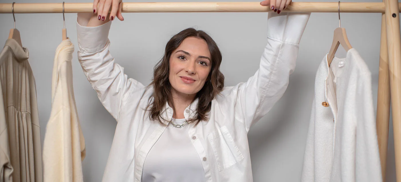 Eine Person in weißer Kleidung streckt sich fröhlich vor einer Kleiderstange mit hellen Kleidungsstücken.
