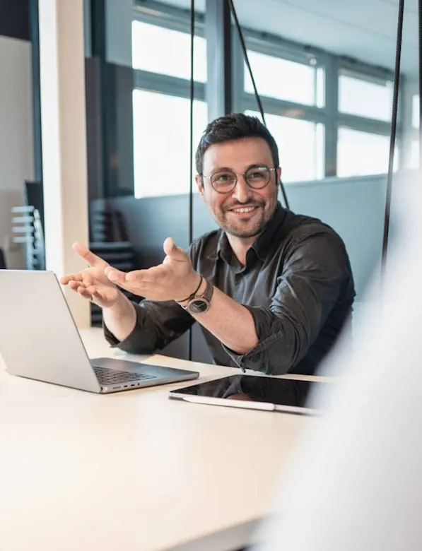 Mann mit Brille gestikuliert fröhlich am Schreibtisch vor Laptop und Tablet im hellen Büro.