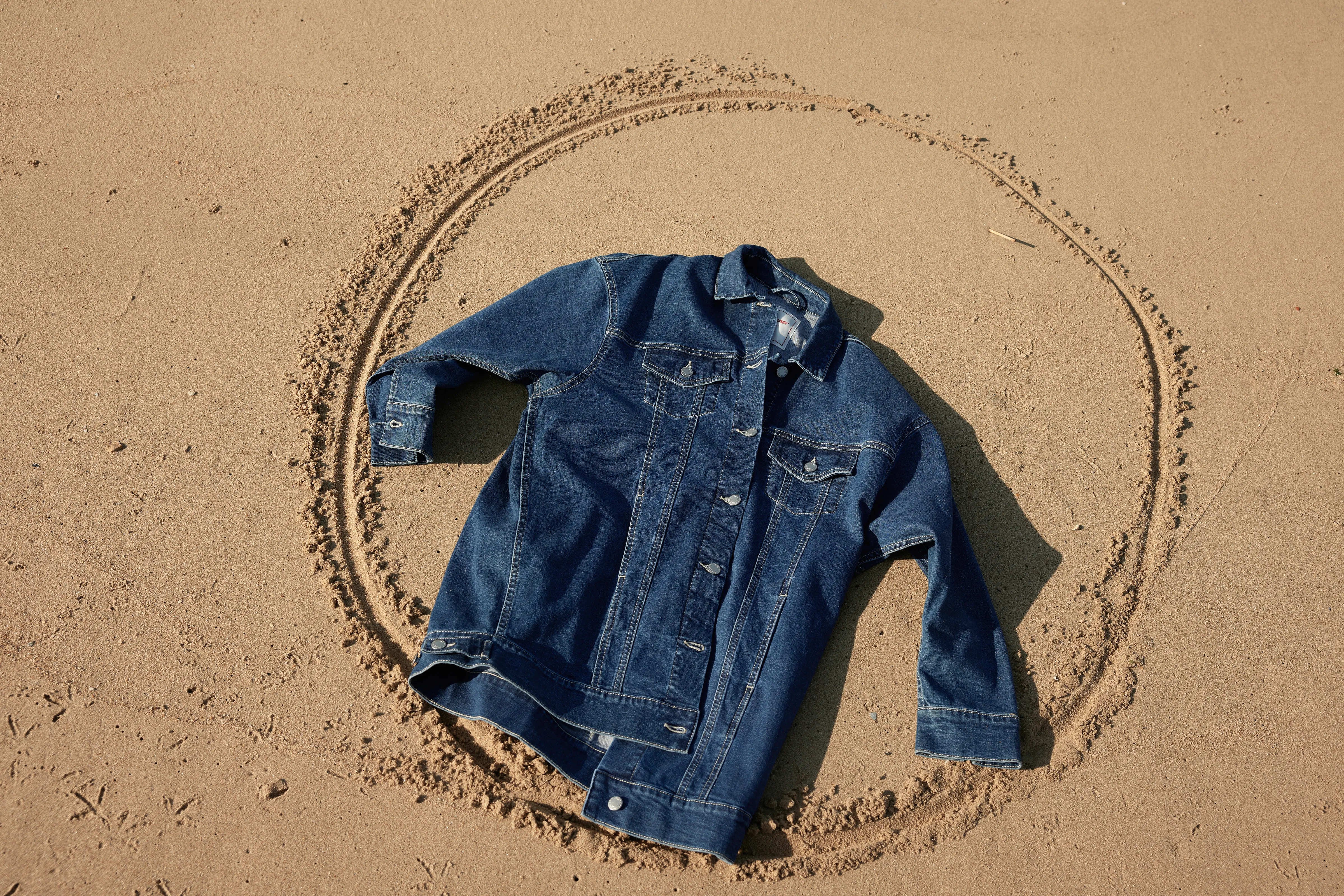 Eine dunkelblaue Jeansjacke liegt auf dem Sand. Um sie herum ist ein Kreis gezeichnet.