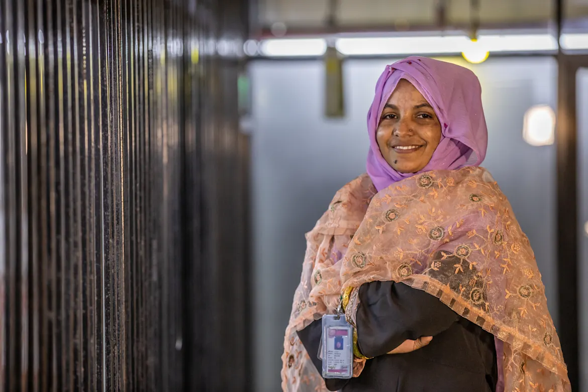 Eine Frau in buntem Hijab lächelt freundlich. Sie trägt ein Namensschild an ihrer Kleidung.